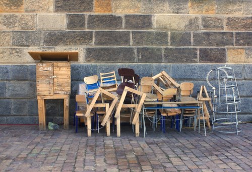A collection of various wooden furniture pieces and metal chairs arranged against a weathered brick wall on a cobblestone surface outside. The furniture includes small chairs, stools, and tables, some with light-colored wooden finishes and others with darker wood tones. There are also metal-framed chairs with blue, silver, and black finishes stacked and leaning against each other. To the left, a wooden cabinet with a sloped top and a small door is positioned directly on the ground; the cabinet appears aged with a rustic appearance. The scene depicts an outdoor area, likely part of a rubbish removal or clearance service in Camden Town, with the furniture awaiting collection or disposal. The backdrop shows a brick wall characteristic of the local urban environment, and the cobblestone pavement suggests a street or alleyway setting.