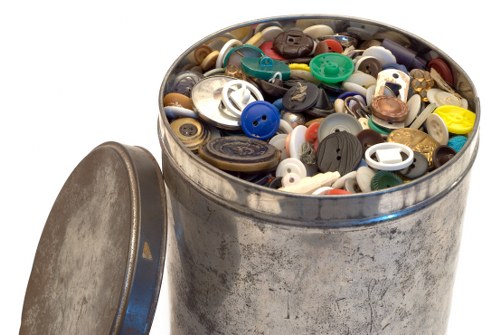 A close-up image of a round, metallic trash can filled to the brim with a variety of plastic buttons in multiple shapes, sizes, and colours, including blue, black, white, yellow, and green. The container has a weathered, slightly tarnished surface and is positioned outdoors on a paved area, with a matching lid resting beside it. The background is plain white, highlighting the focus on the detailed collection of buttons and the container's metallic texture, relevant for waste management services specializing in rubbish clearance, particularly in Camden Town or surrounding areas.