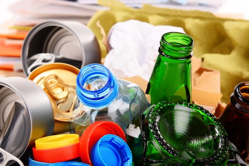 A collection of assorted glass bottles and jars, including green, brown, and clear varieties, some with metal caps or no lids, are scattered on a surface alongside plastic lids in red, yellow, blue, and orange colors, as well as crumpled paper and cardboard packaging. The bottles and lids are of different shapes and sizes, with some appearing used or discarded, indicative of waste materials awaiting collection or removal. In the background, partially visible, are grey plastic containers and packaging materials, suggesting a setting related to rubbish or waste disposal, possibly in an outdoor or warehouse environment in Camden Town, London. The natural lighting emphasizes the textures and transparency of the glass, with some reflections visible on their surfaces, highlighting the importance of correct waste segregation for recycling and waste management services provided by Camden Town House Clearance.