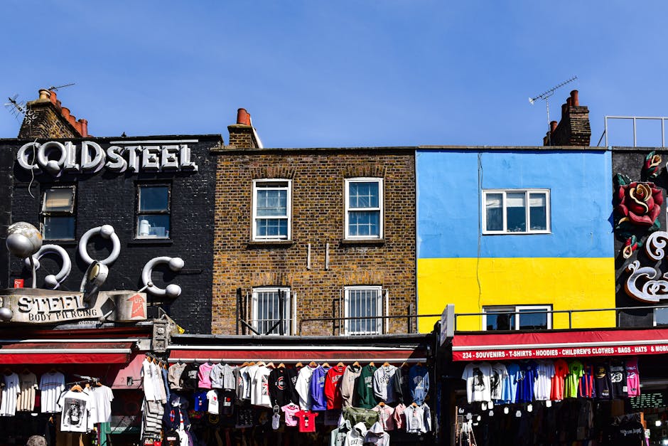 An urban street scene featuring a variety of building facades under a clear blue sky. The leftmost building has a dark brick exterior with a black painted wall, displaying a large metallic sign readin